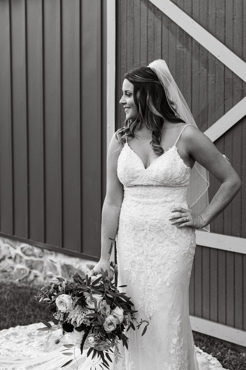 Monochrome side view of bride in lace gown holding bouquet against barn wall.