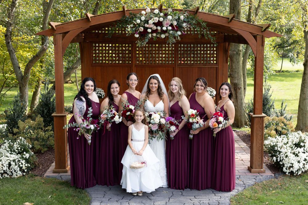 Bridal party in burgundy dresses gathered under wooden gazebo decorated with white flowers.