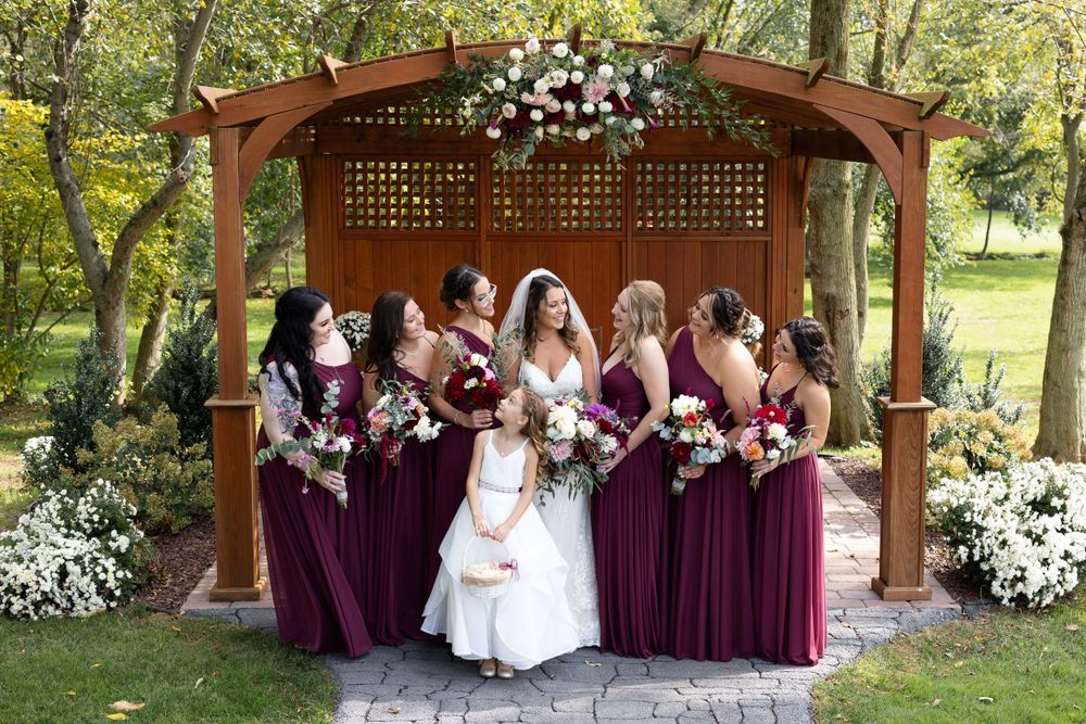 Wedding party poses under floral-decorated wooden pergola in garden setting.