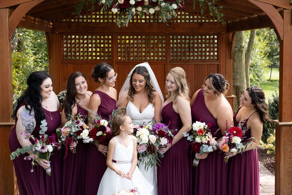 Wedding party sharing joyful moment under wooden pergola with burgundy bridesmaids dresses.