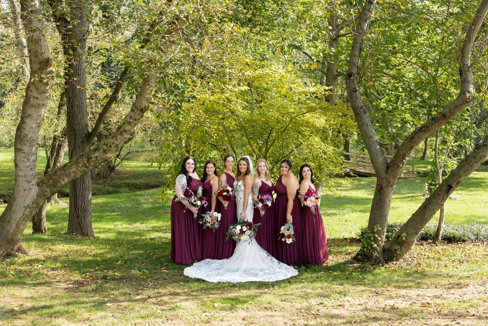 Bridal group in burgundy dresses arranged outdoors among trees in natural setting.