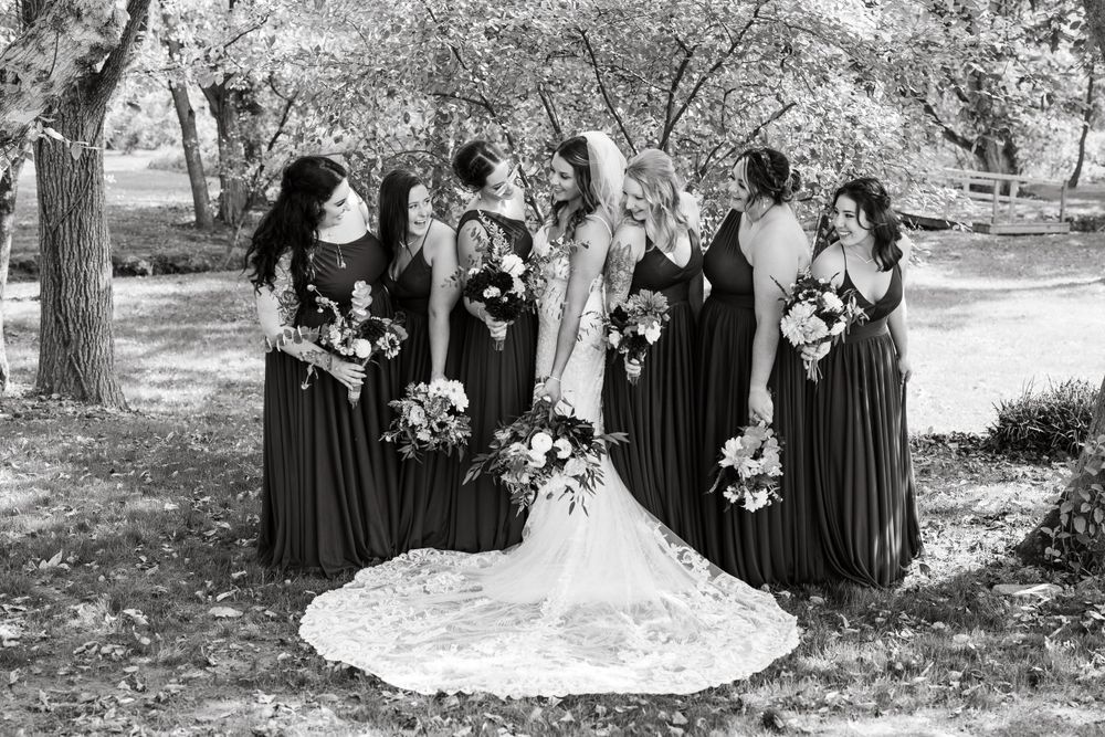Wedding party in black bridesmaid dresses pose with bouquets under trees in a natural outdoor setting.