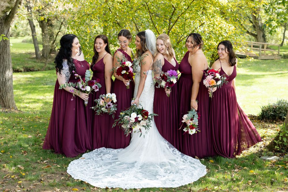 Bridal party in burgundy dresses gather outdoors holding floral bouquets with bride in white lace gown.