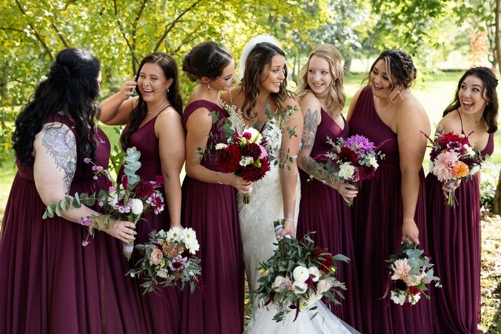 Candid moment of bridal party in burgundy dresses holding bouquets and laughing together outdoors.