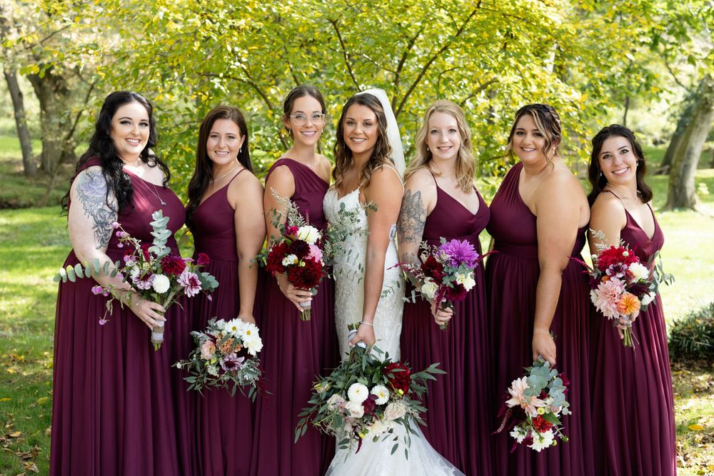 Wedding party in burgundy dresses pose with bouquets against a backdrop of autumn trees.