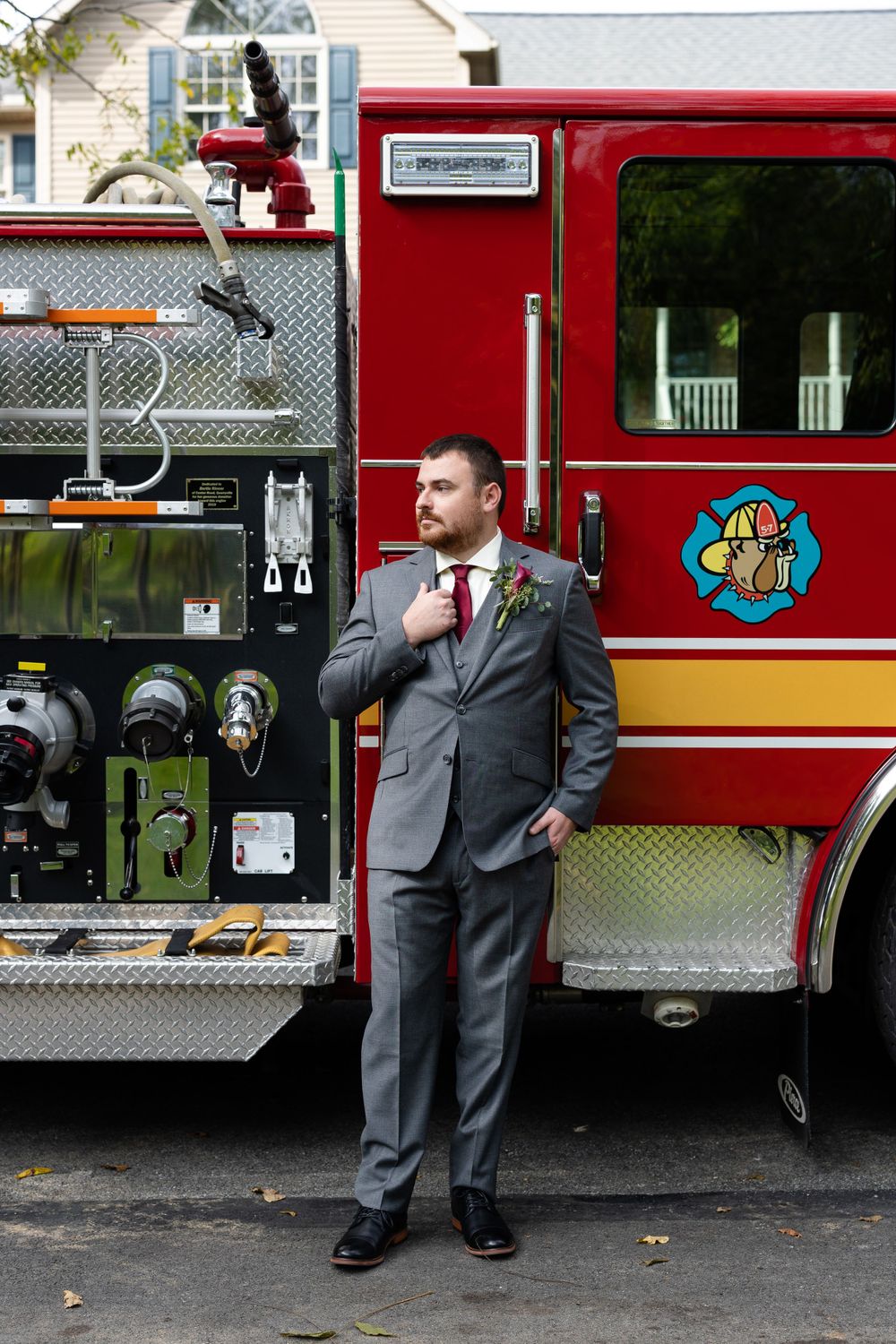 Groom in grey suit poses in front of red fire truck wearing burgundy tie and boutonniere.