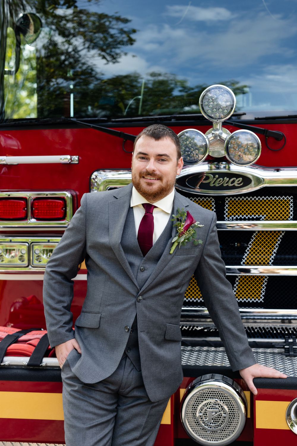 Groom in grey suit poses in front of vintage fire truck with chrome details.