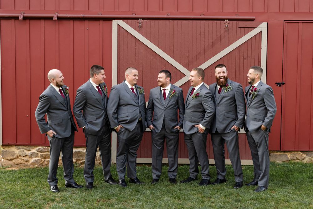 Wedding party members in matching grey suits pose in front of a red barn door.