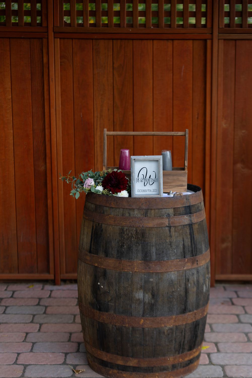 Rustic wooden barrel decorated with wedding decor and signage on brick paving.