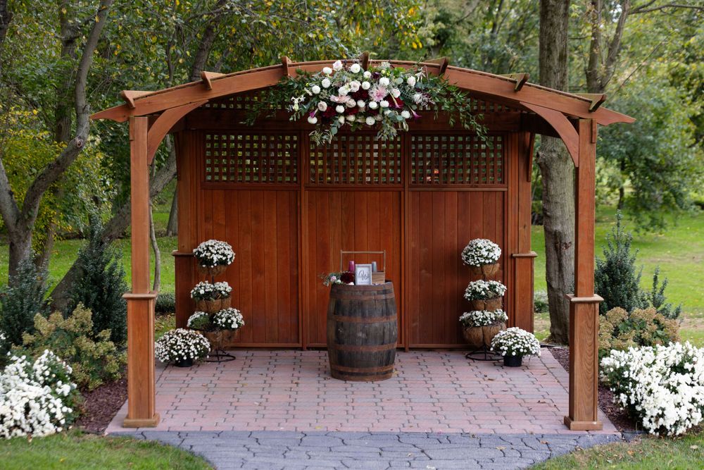 Wedding ceremony setup with wooden pergola, barrel, and floral arrangements in garden setting.