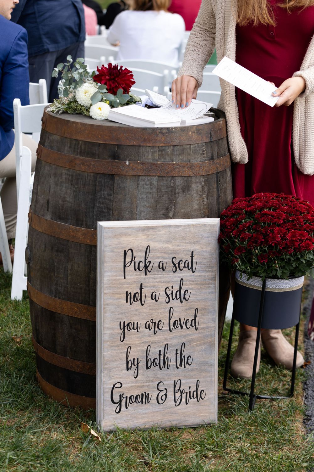 Rustic wooden barrel with wedding seating sign and red flowers at outdoor ceremony.