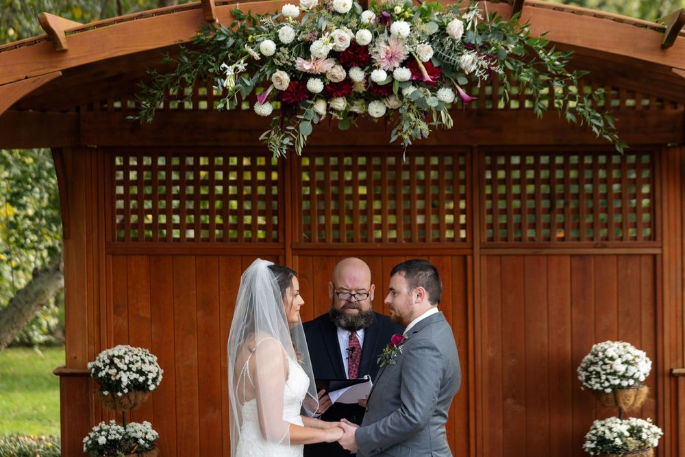 Wedding ceremony taking place under wooden pergola decorated with white and burgundy floral arrangements.