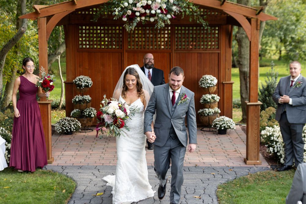 Newlyweds exit wooden gazebo decorated with white flowers and burgundy accents.