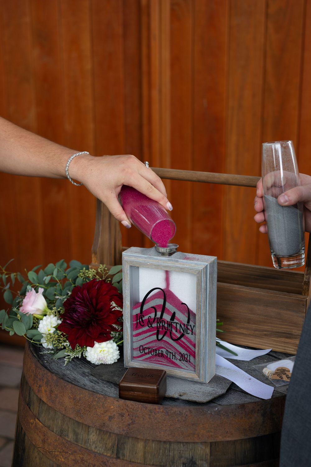 Pouring colored sand into decorative frame at rustic barrel wedding ceremony display.