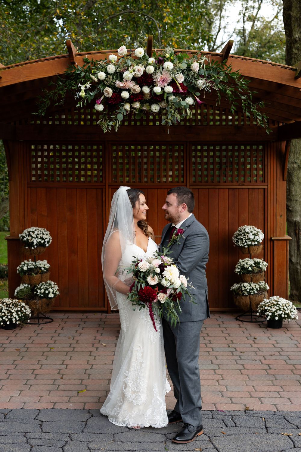 Elegant couple poses under floral-adorned wooden pergola with burgundy and white bouquet.