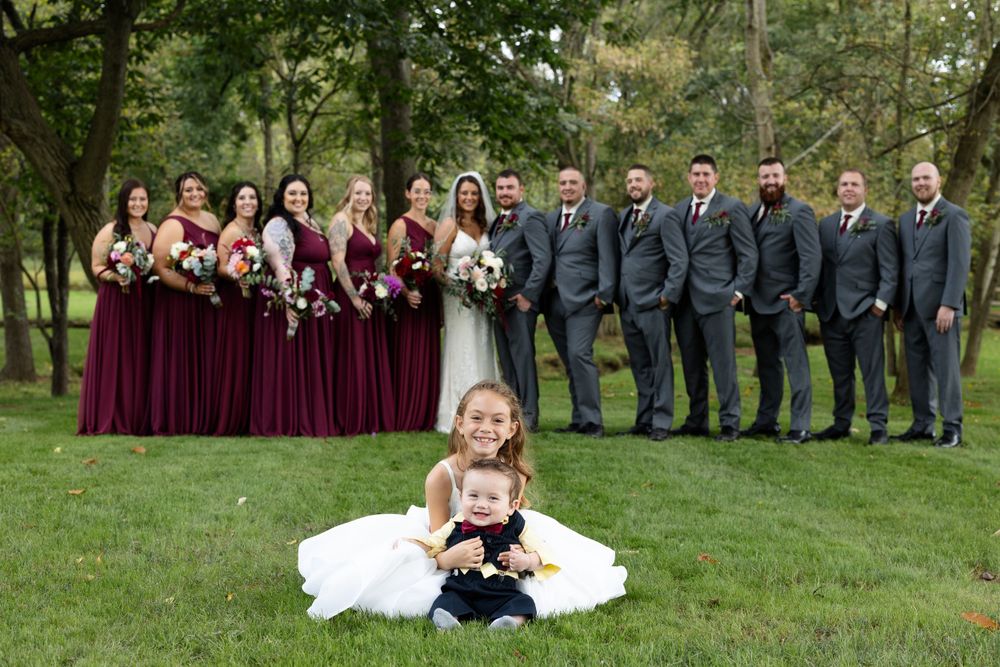 Wedding party in burgundy dresses and grey suits pose with flower children on grass.