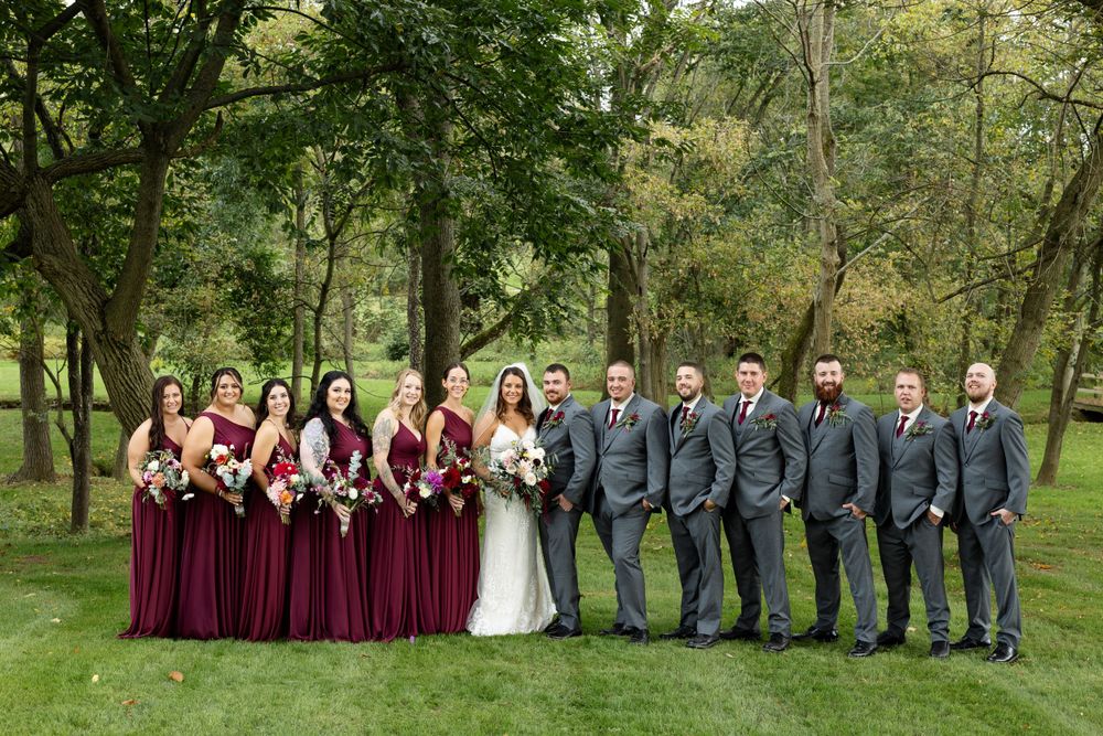 Formal wedding party lineup with burgundy bridesmaids and groomsmen in grey suits outdoors.