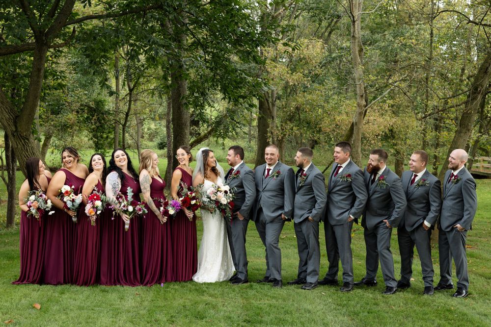 Wedding party stands in line on green grass wearing burgundy dresses and gray suits in outdoor setting.