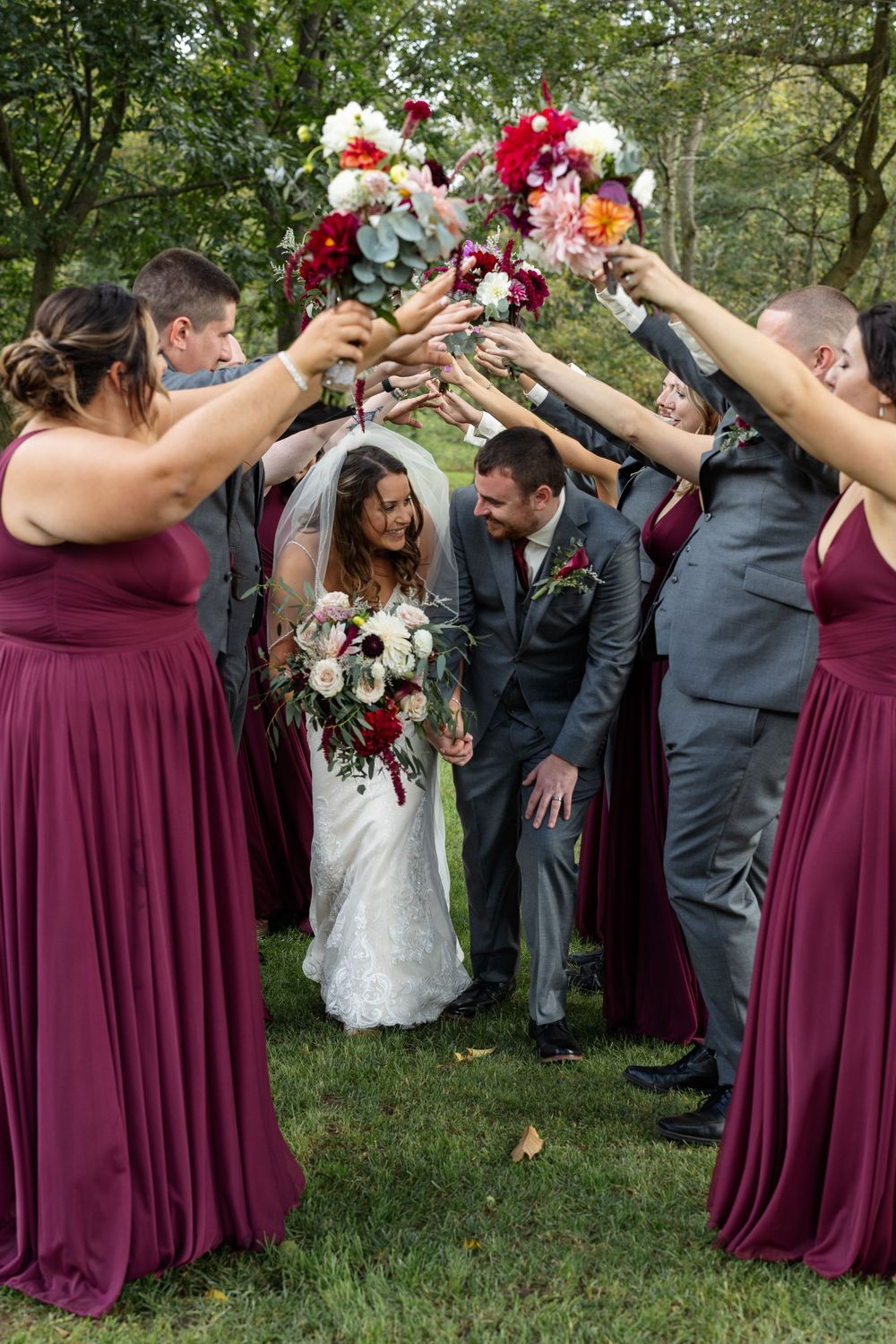 Wedding party forms flower arch tunnel for couple in burgundy and gray attire.