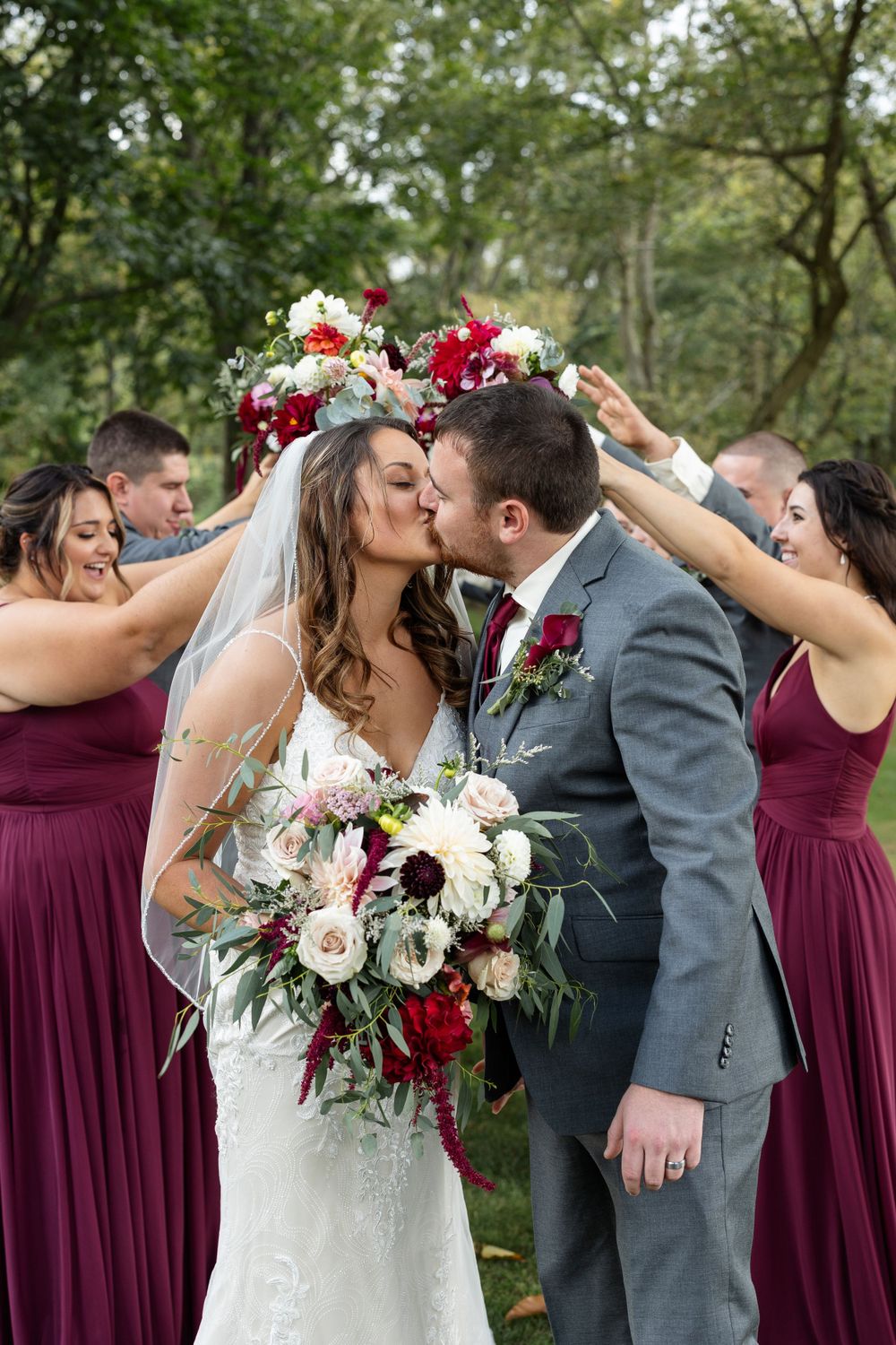 Couple shares romantic moment surrounded by burgundy-dressed wedding party with red and white bouquet.