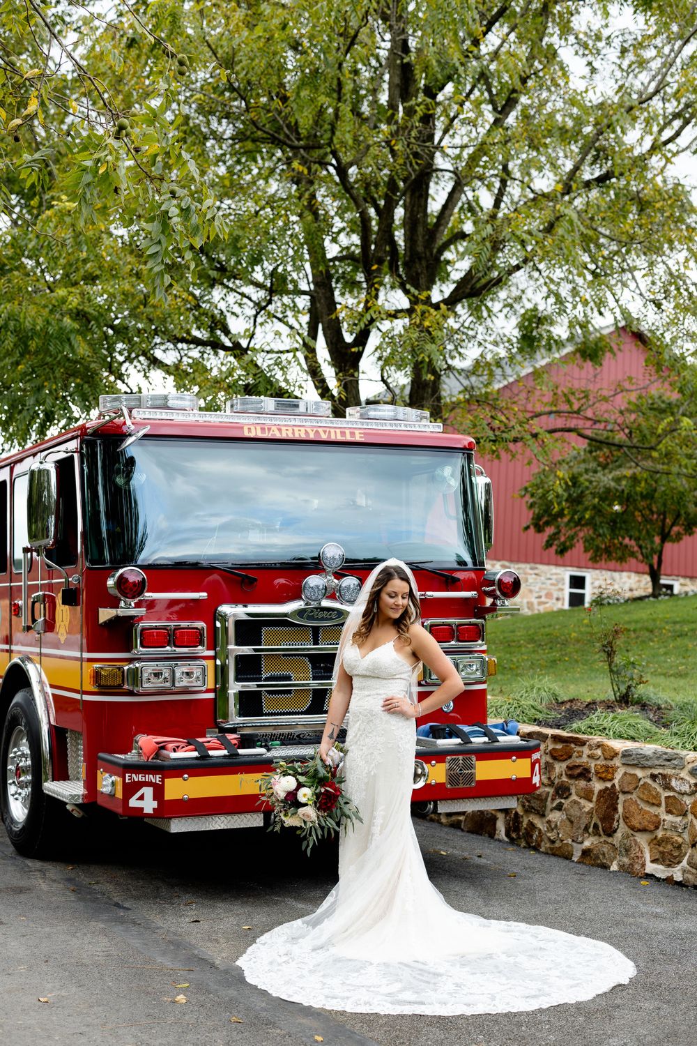 A bride in a flowing white dress poses beside a red fire engine with fall foliage in the background.