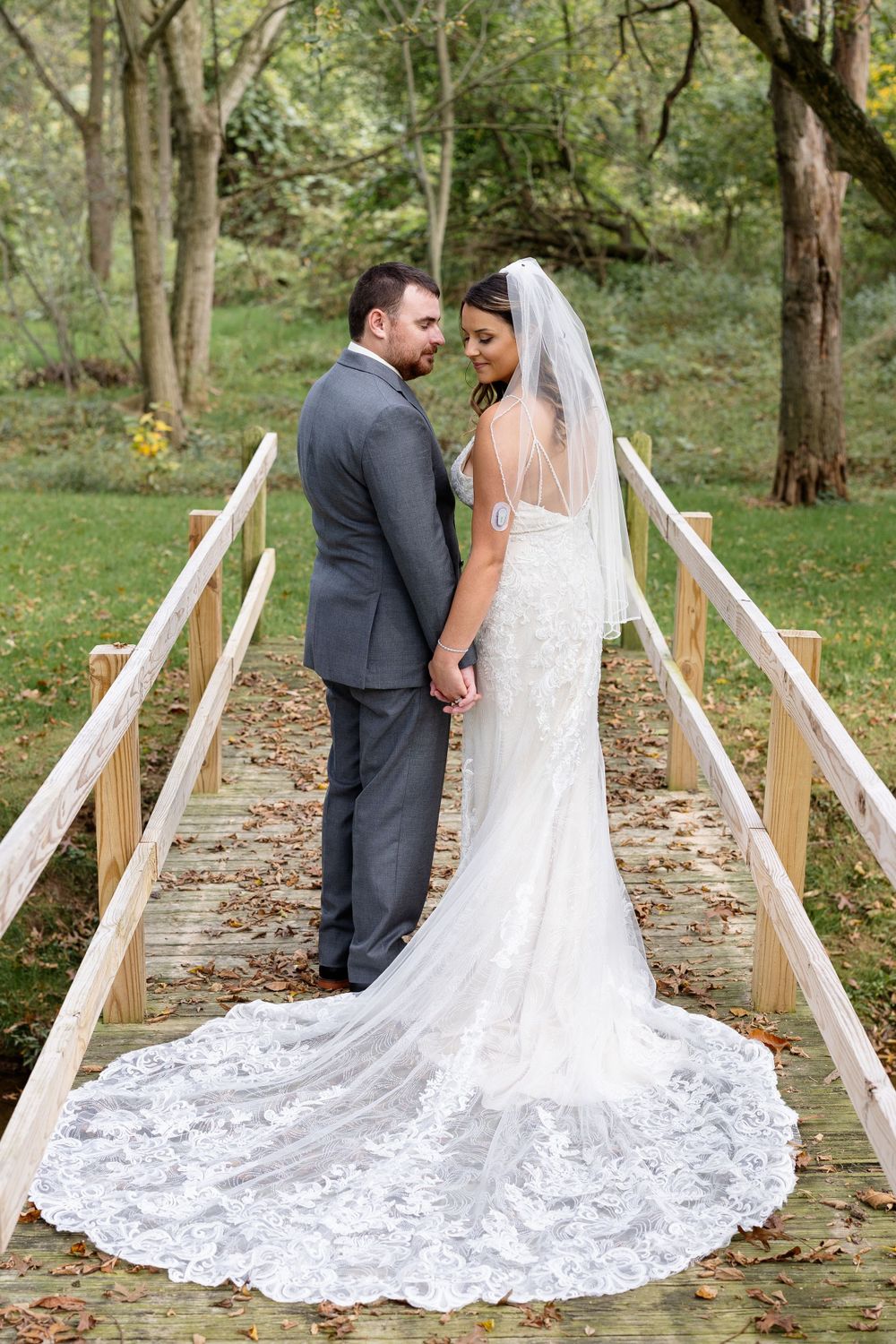 Couple in wedding attire standing on wooden bridge with flowing lace train.