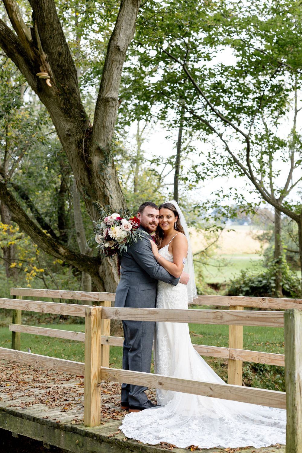 Newlyweds embrace on rustic wooden bridge with flowering trees in background.