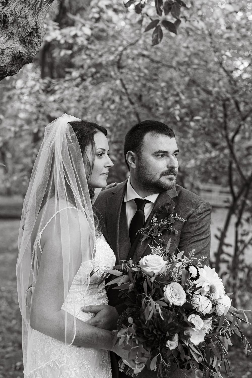 A black and white wedding portrait shows a couple posing outdoors with the bride holding a lush floral bouquet.