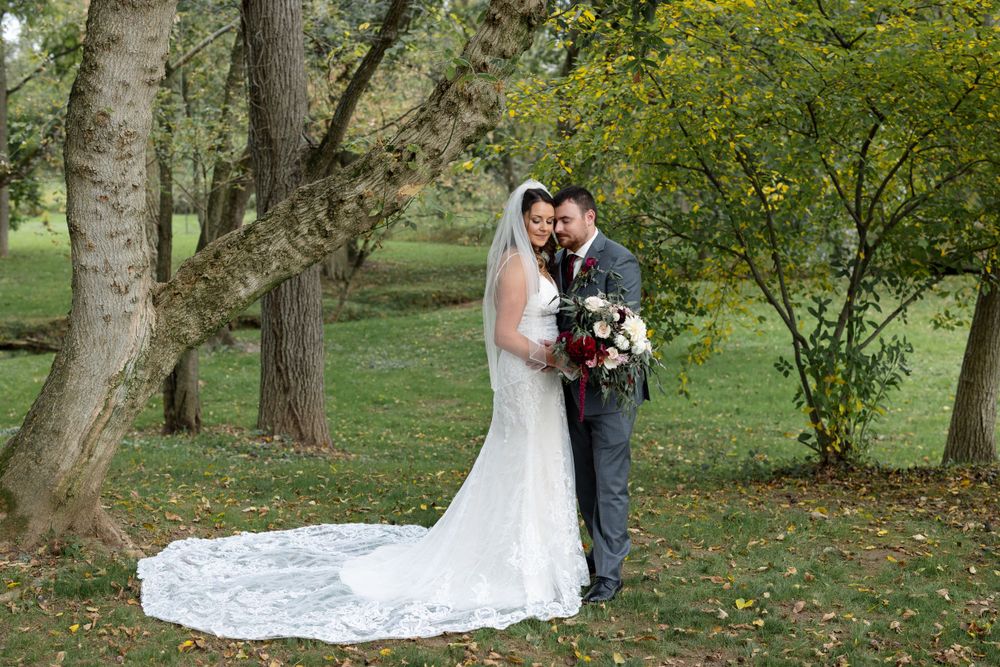 Newlyweds pose together under autumn trees with the bride's long lace train trailing behind.