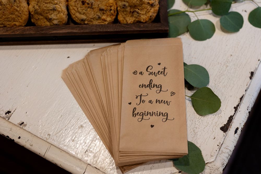 Brown paper wedding favor bags with text 'a sweet ending to a new beginning' arranged with eucalyptus leaves.