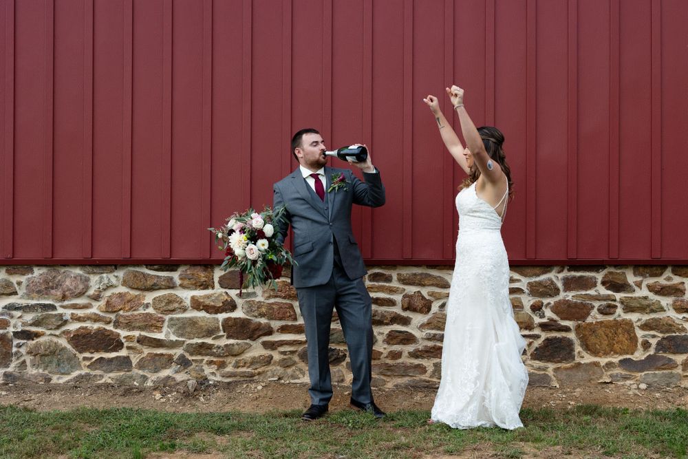 A couple celebrating their wedding day with joyful poses against a rustic red barn wall.