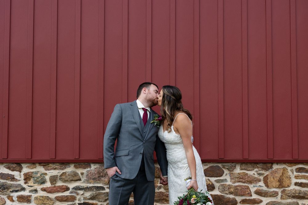 Romantic moment shared between newlyweds against a red barn with stone foundation.