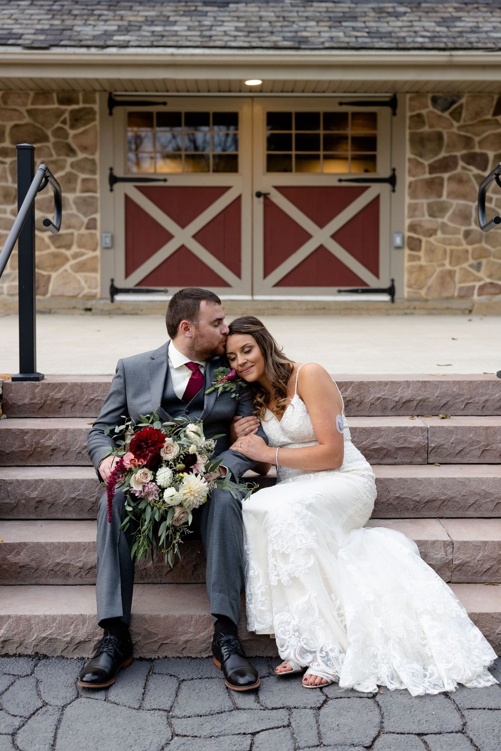 Newlyweds sharing an intimate moment on stone steps with rustic barn doors in background.