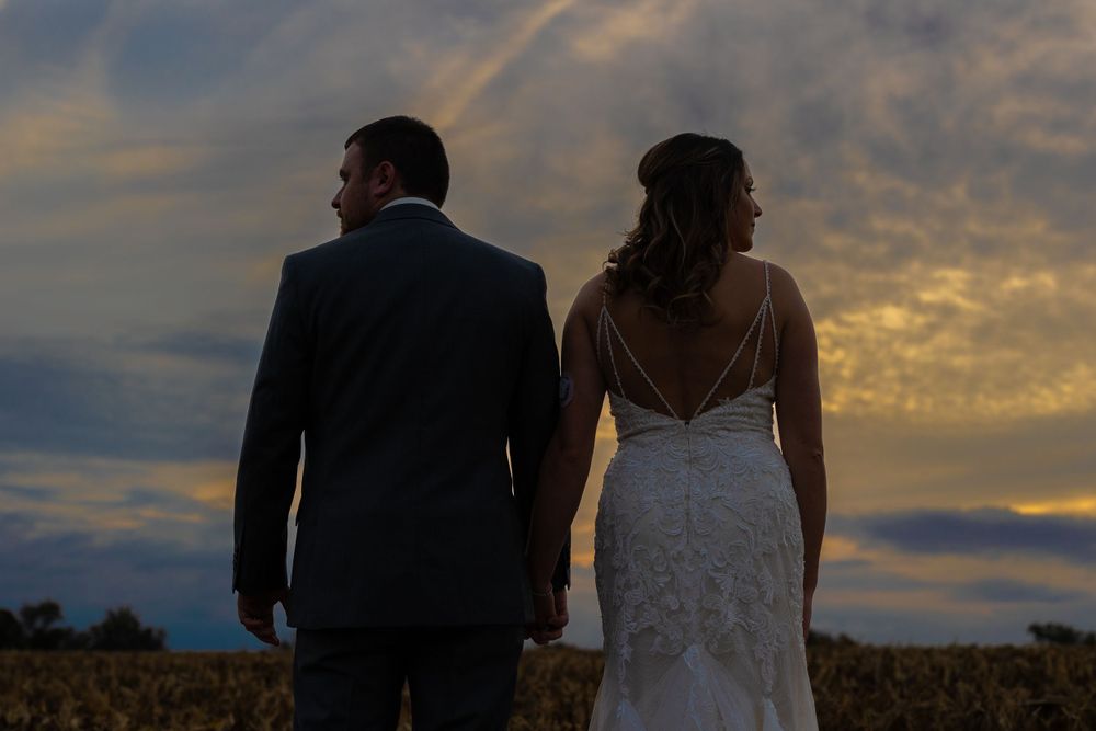 Newlyweds stand together against a dramatic sunset sky in an autumn field.
