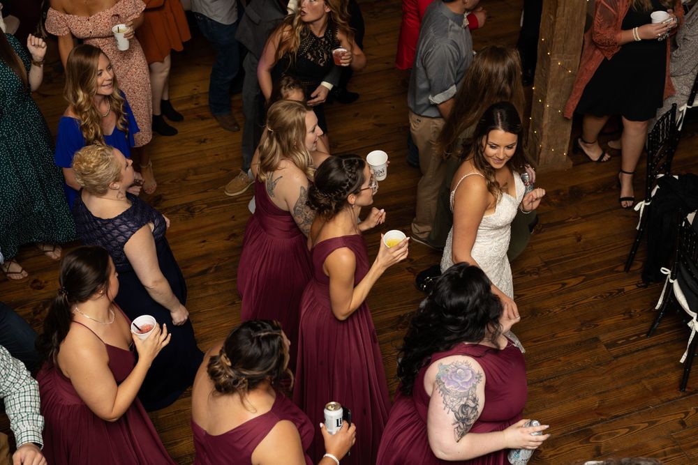Wedding party celebrates with dancing and drinks at a rustic indoor reception venue.