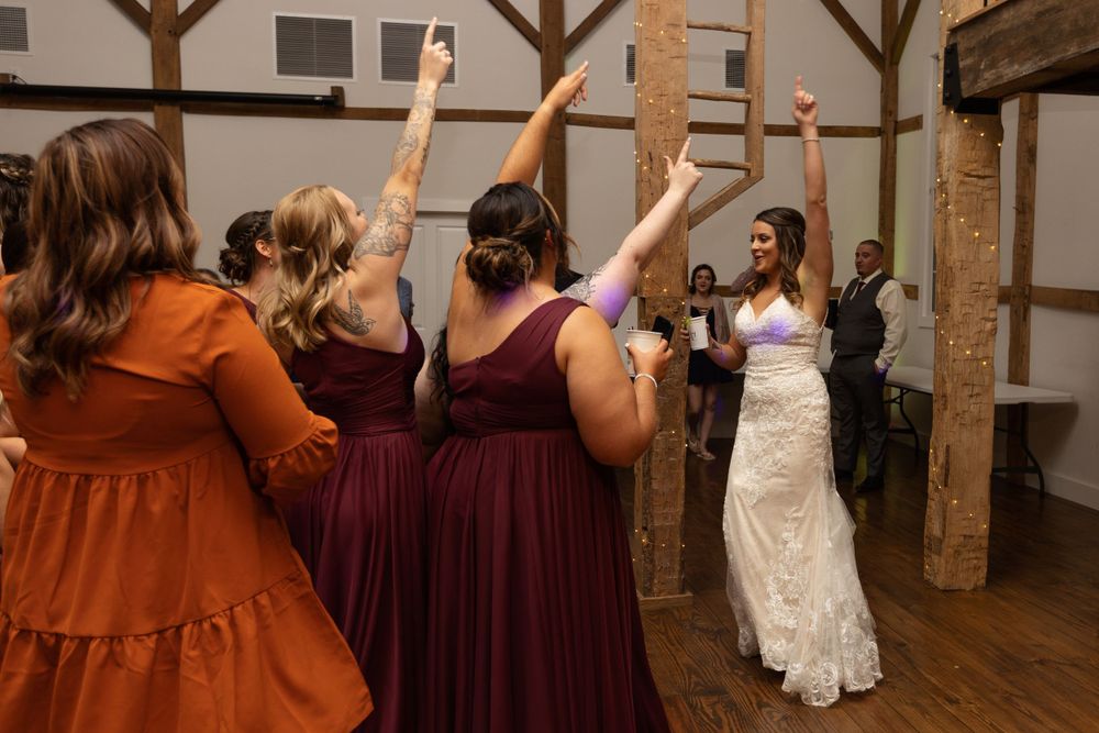 Wedding attendees in burgundy and rust colored dresses dance with the bride in a rustic venue.