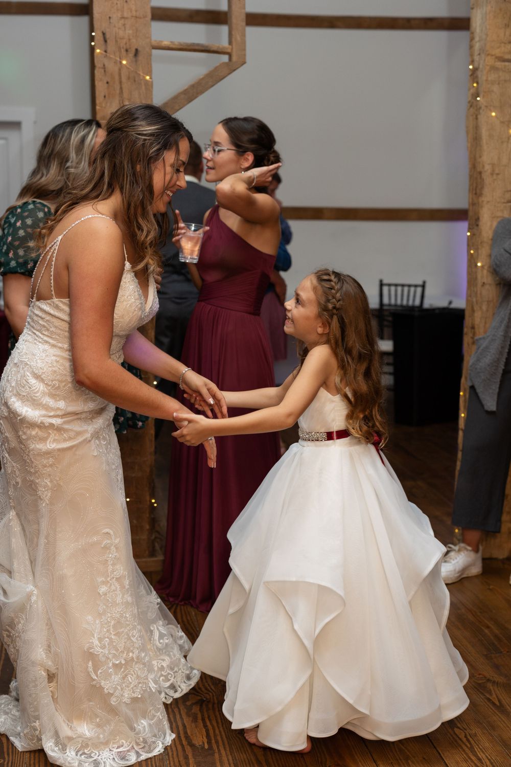 A white wedding dress adorned with lace dances with a young flower girl in a layered white dress at a reception.