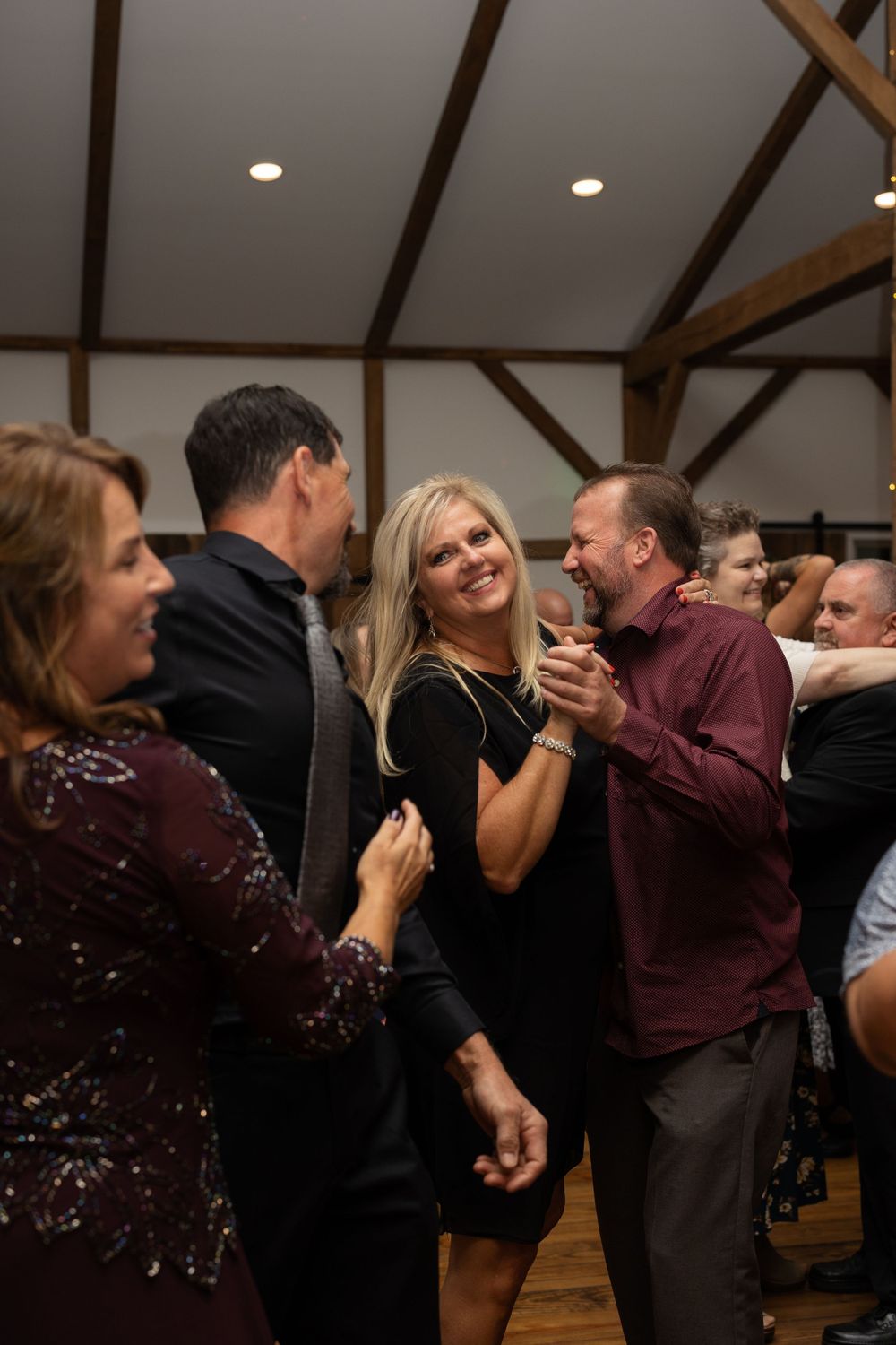 Wedding guests dance and laugh together on a wooden dance floor under exposed wooden beams.