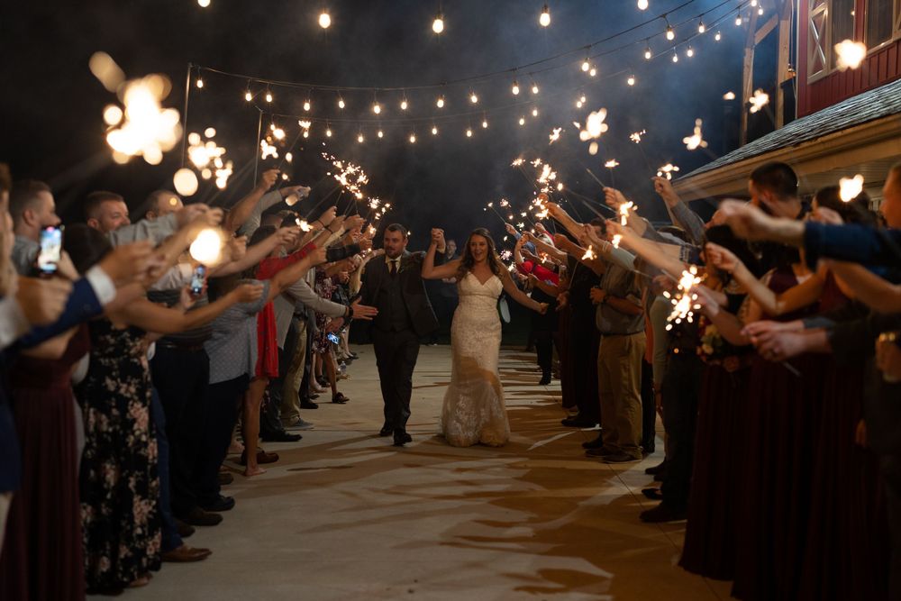 Wedding couple exits through sparkler sendoff line under string lights at night.