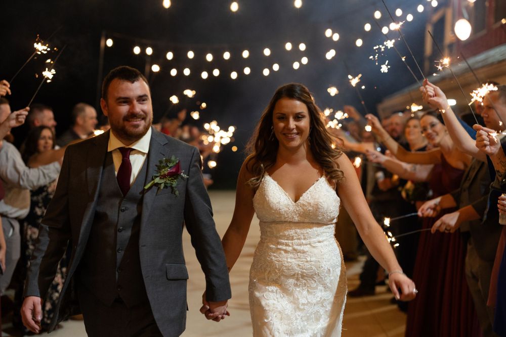Couple walks down the aisle at their wedding reception under twinkling string lights while guests celebrate.