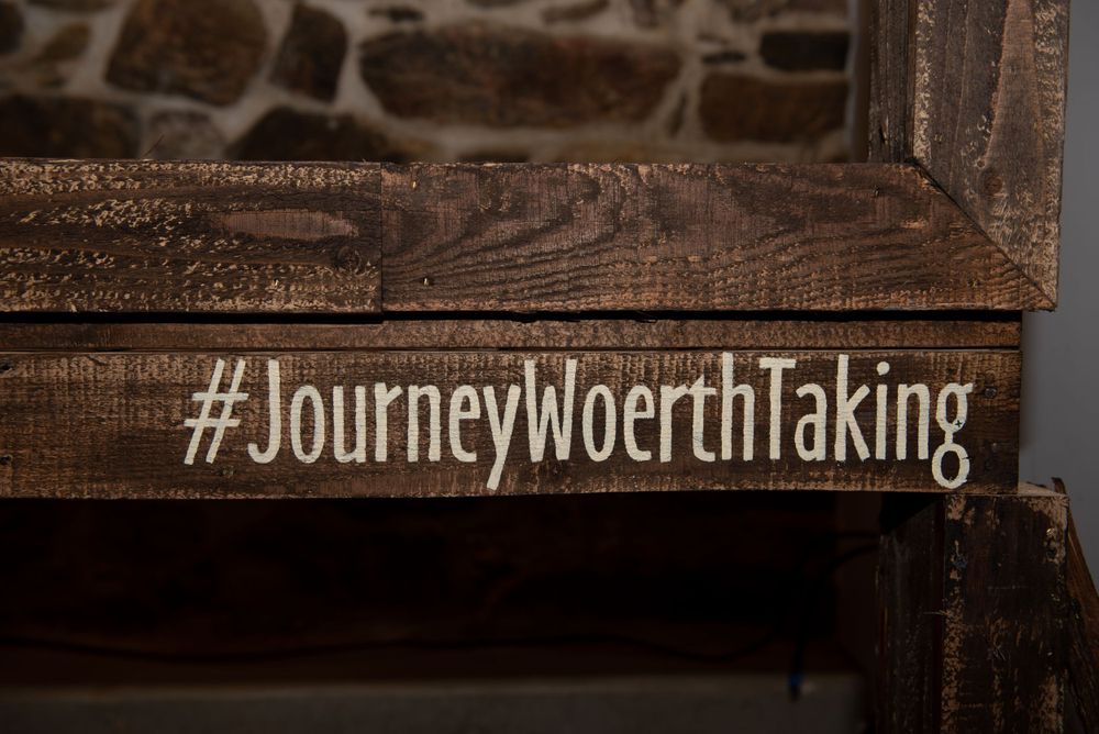 Wooden sign with hashtag JourneyMoerthTaking engraved against rustic stone wall background.