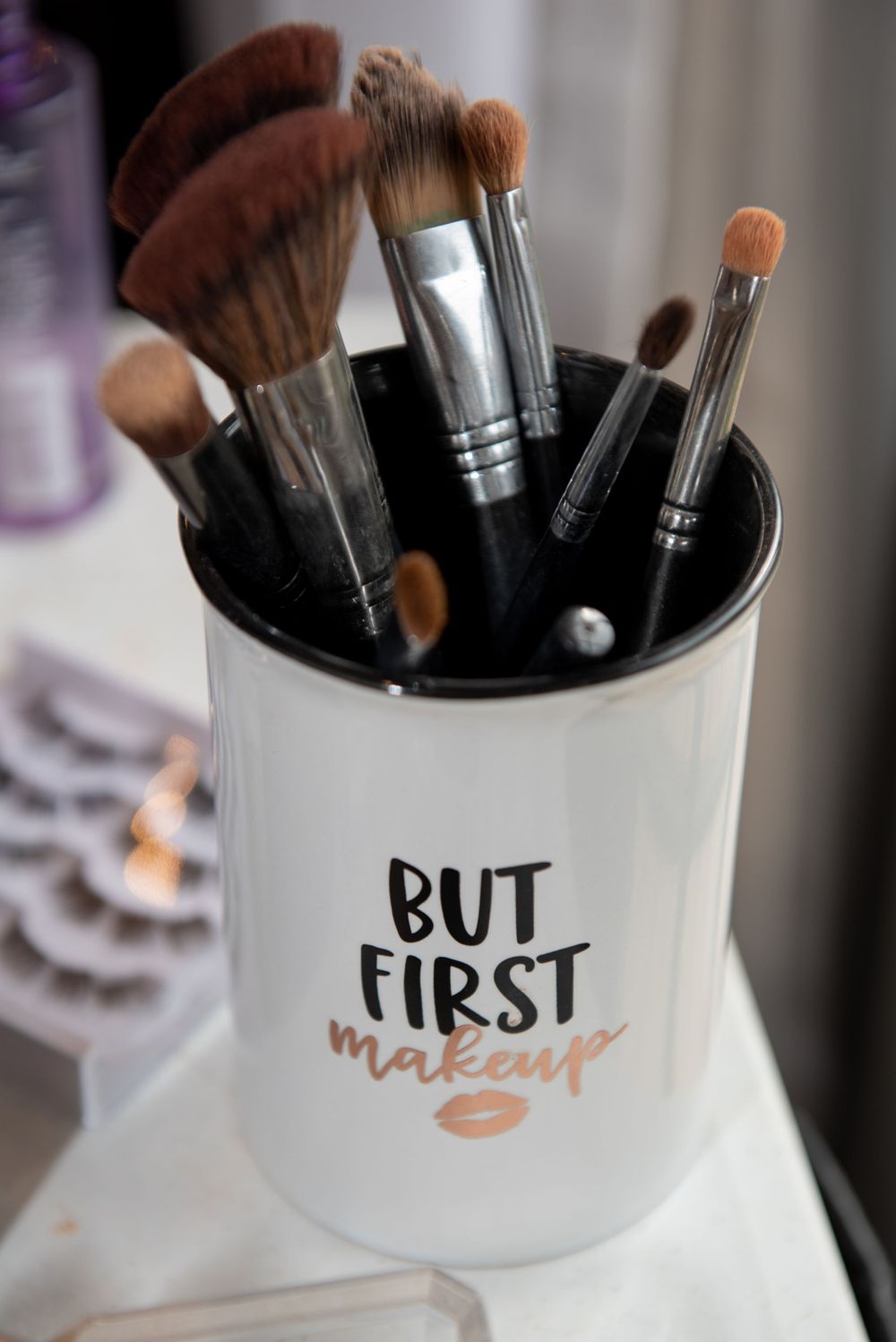 Makeup brushes displayed in white ceramic holder with 'But First Makeup' text and kiss mark design.
