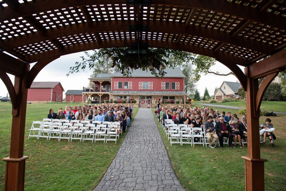 Outdoor wedding ceremony setup with white chairs under a wooden pergola facing a red barn.