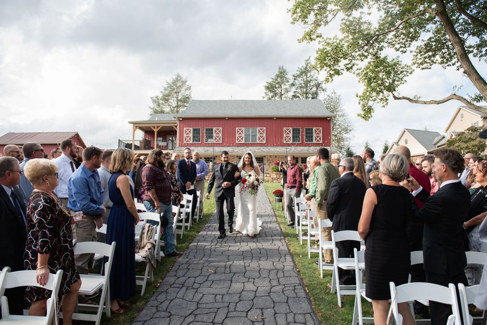 Wedding ceremony procession on a stone pathway with guests seated on white chairs.