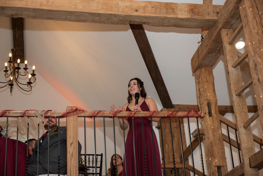 A woman in a burgundy dress stands at a rustic wooden balcony with exposed beams and chandelier.