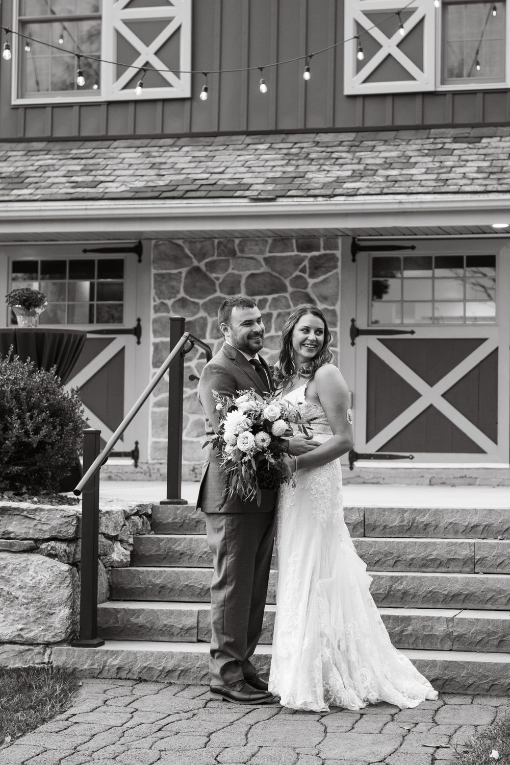 A couple poses together on stone steps outside a barn-style building with white X-framed details.