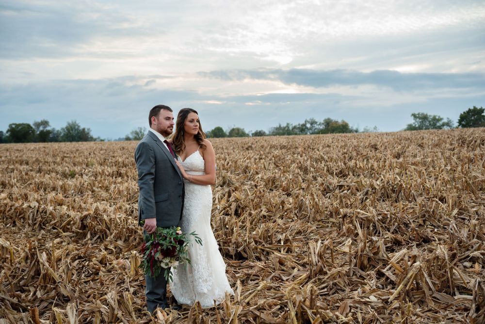 A couple stands together in a harvested cornfield at sunset against a cloudy sky.