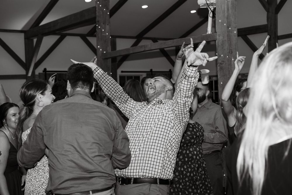 People dancing and celebrating at a wedding reception with exposed wooden beams overhead.