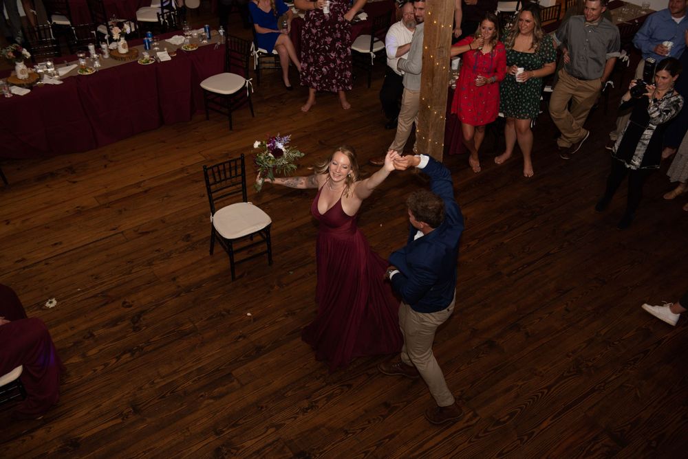 People dancing together in a rustic reception venue with wooden floors and banquet tables.