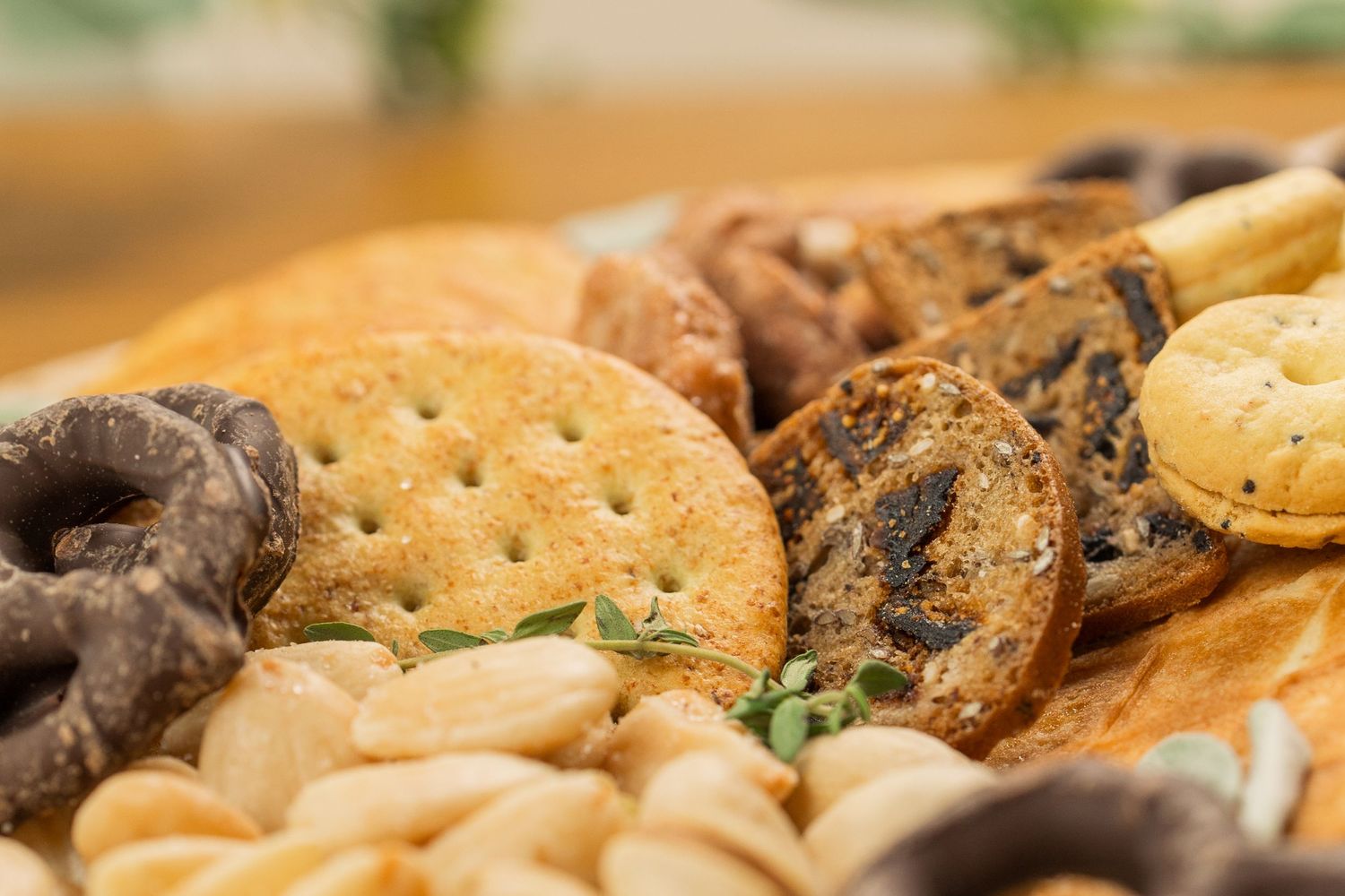 Close-up of a snack platter with crackers, chocolate-covered pretzels, and nuts garnished with fresh herbs.